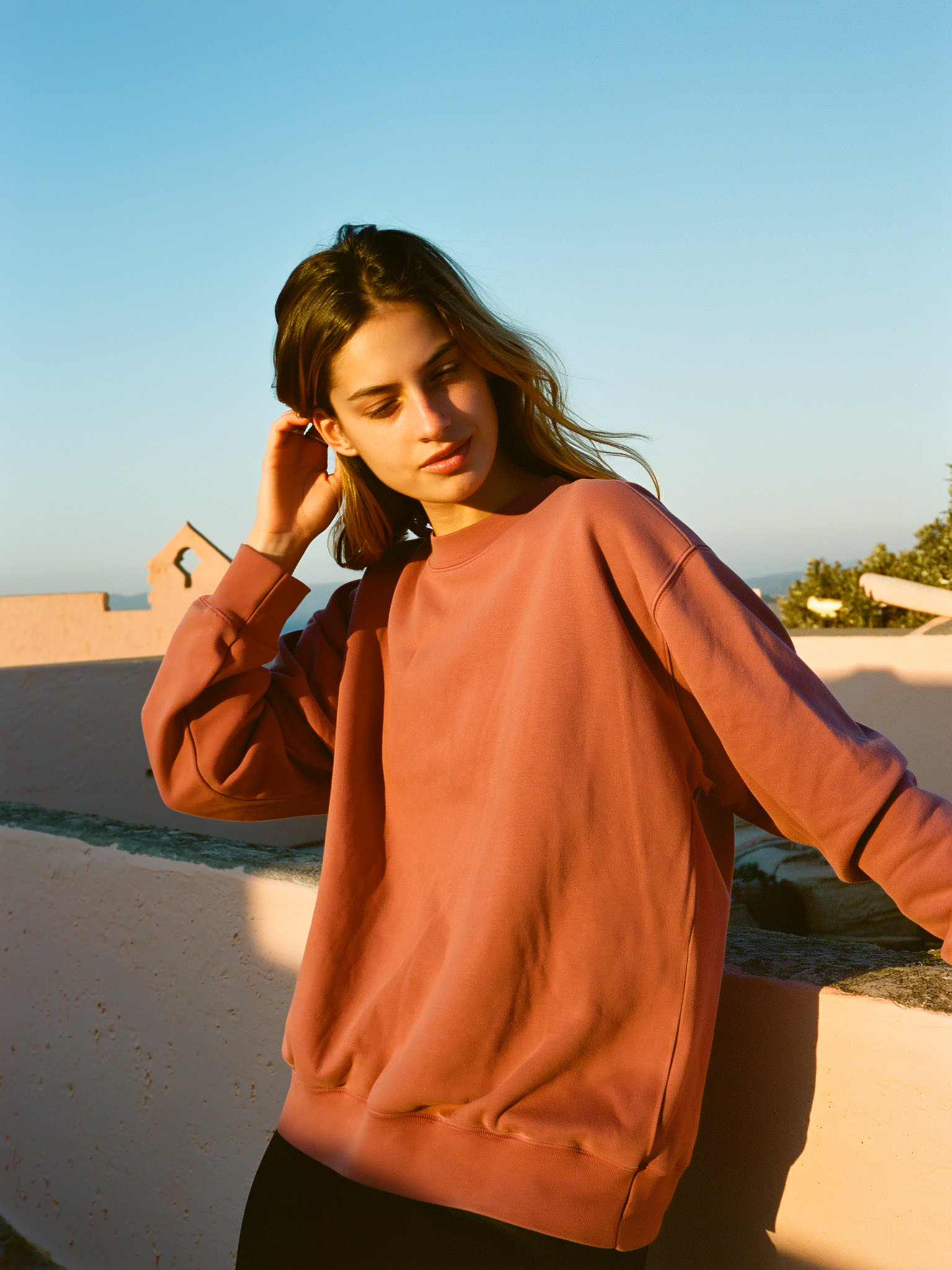 Model in a dusty terracotta heavyweight crewneck against a warm plaster wall